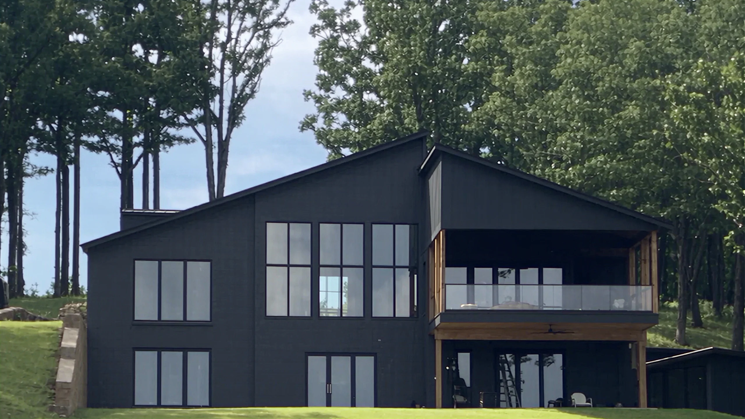Architectural visualization of a lakefront custom home: a dark two-story house with tall windows and a timber balcony above a sloping lawn.